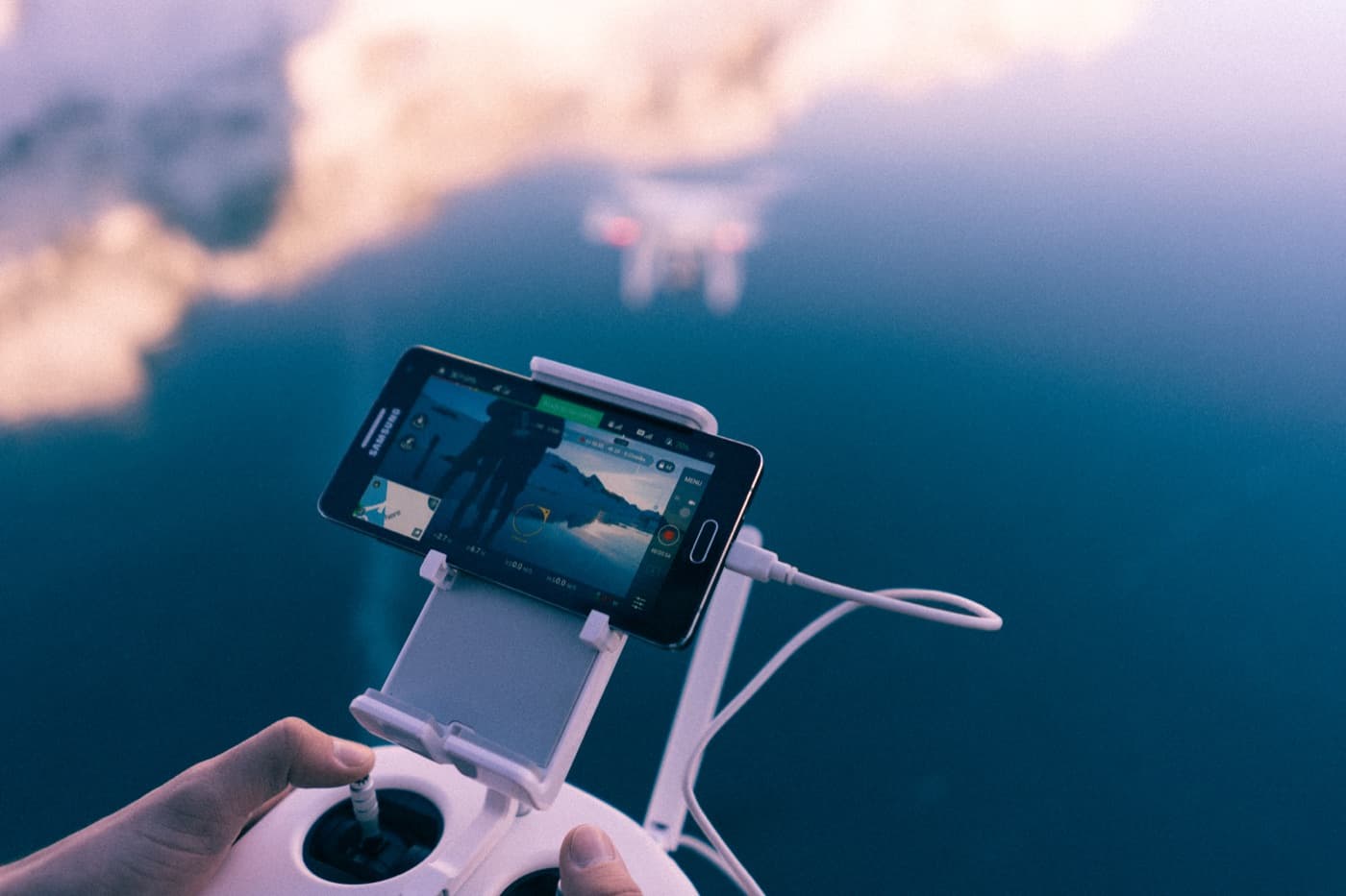 Hands on a drone controller with a phone showing the camera view, the drone hovering over water